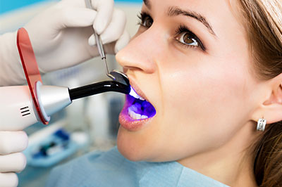 A woman receiving dental care with a dental mirror and light, in the foreground, while wearing a surgical mask.