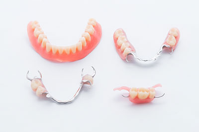 The image displays a collection of dentures and dental appliances, including upper and lower sets with teeth, gums, and metal clasps or attachments, arranged on a white background.
