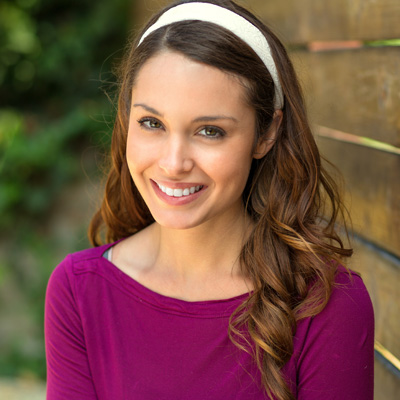 A smiling woman with long brown hair, wearing a purple top and a headband, poses against a wooden fence.