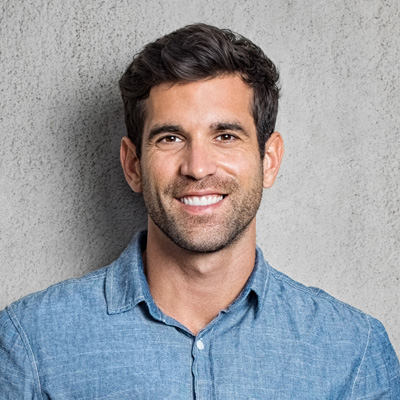 A man with a beard is smiling and looking directly at the camera. He has short hair, is wearing a blue denim shirt, and stands against a gray background.