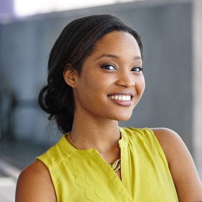 The image features a young woman with a bright smile, wearing a yellow top and standing against a concrete wall.