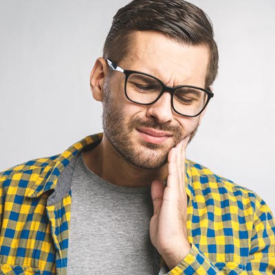 The image shows a man with glasses, wearing a plaid shirt, holding his hand to his mouth as if he is in pain or discomfort.